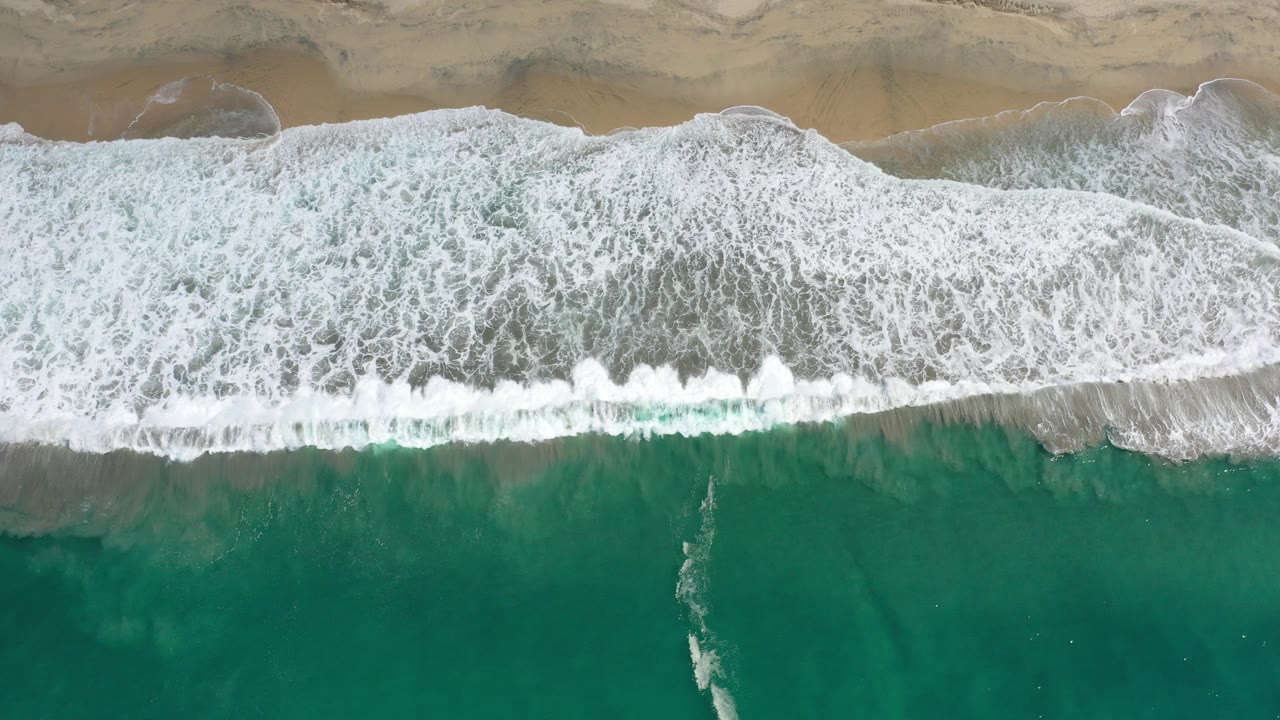 Overhead view in Los Cabos, Mexico, with green water below, a wide band of white surf in the center, tan sand at the top, and no visible people.