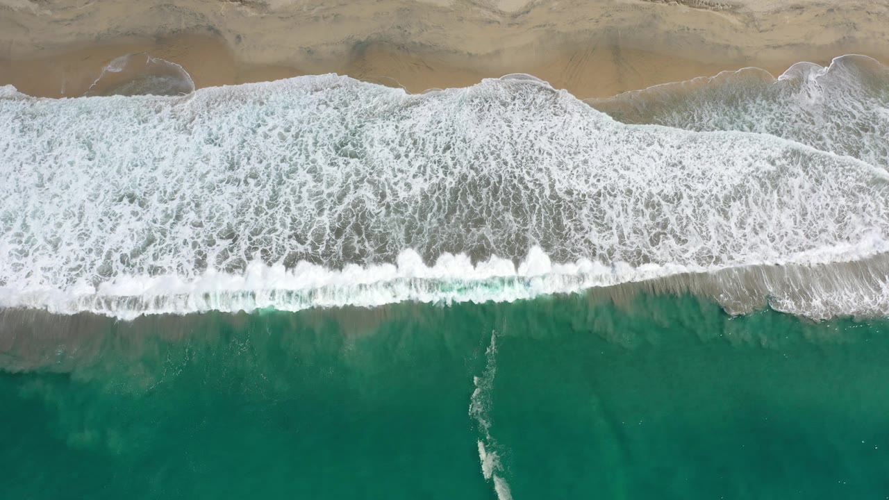 Aerial Los Cabos shoreline frame with foamy surf spread farther across the center and green water still visible below, with no visible people.