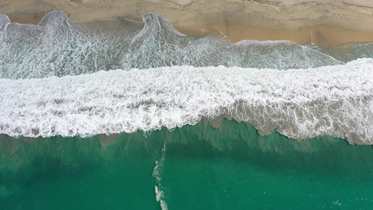Overhead view in Los Cabos, Mexico, with white surf crossing the center of the frame, pale sand above, green water below, and no visible people.