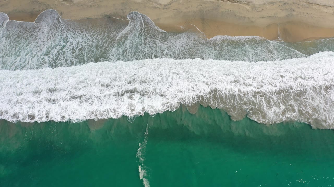Top-down Los Cabos beach frame with the foamy break spread wider across the middle, tan sand at the top, green water at the bottom, and no visible people.