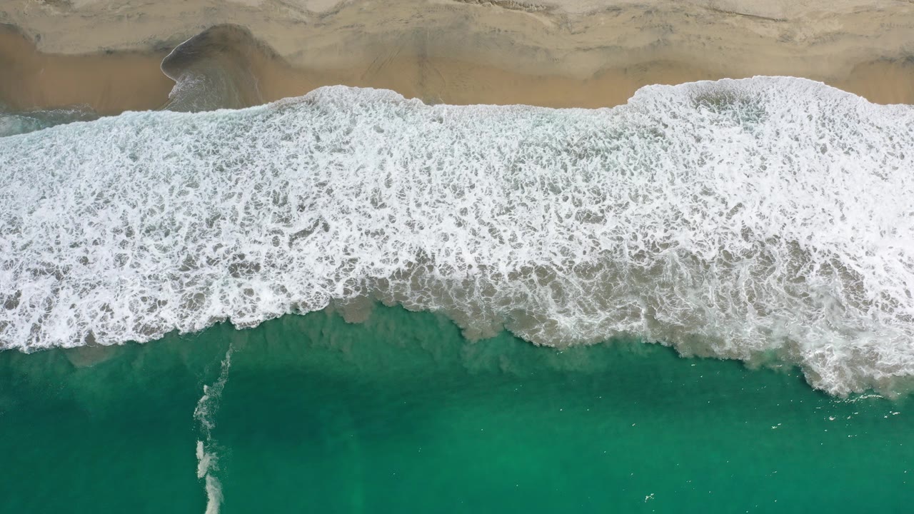Overhead view in Los Cabos, Mexico, with white surf pressing across tan sand and green water below, no visible people.