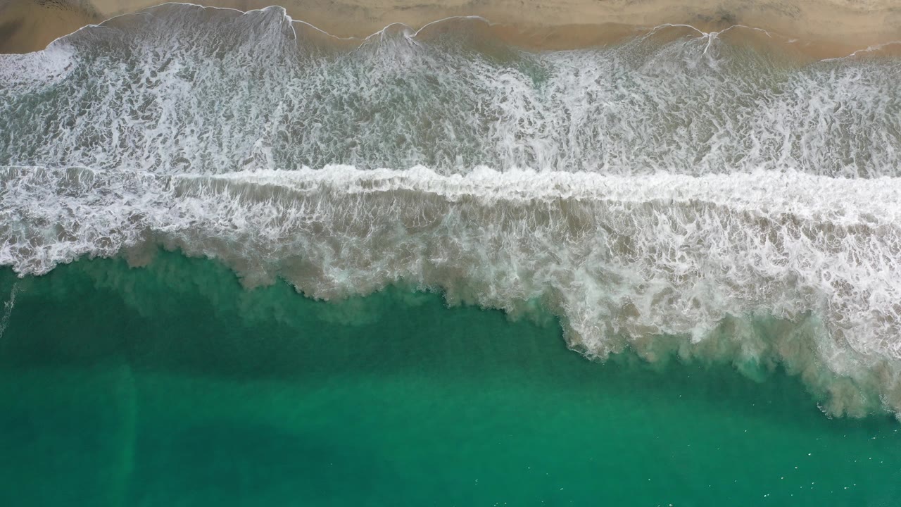 Overhead view of Los Cabos surf at the start of the clip, with white foam spread across teal water below tan beach and no visible people.