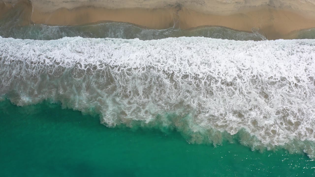 Overhead view of green water and white surf reaching a tan Los Cabos beach at the start of the clip, with no visible people.
