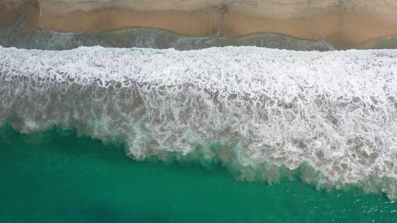White wave foam spreads farther across green water below a thin strip of Los Cabos sand, with no visible people.