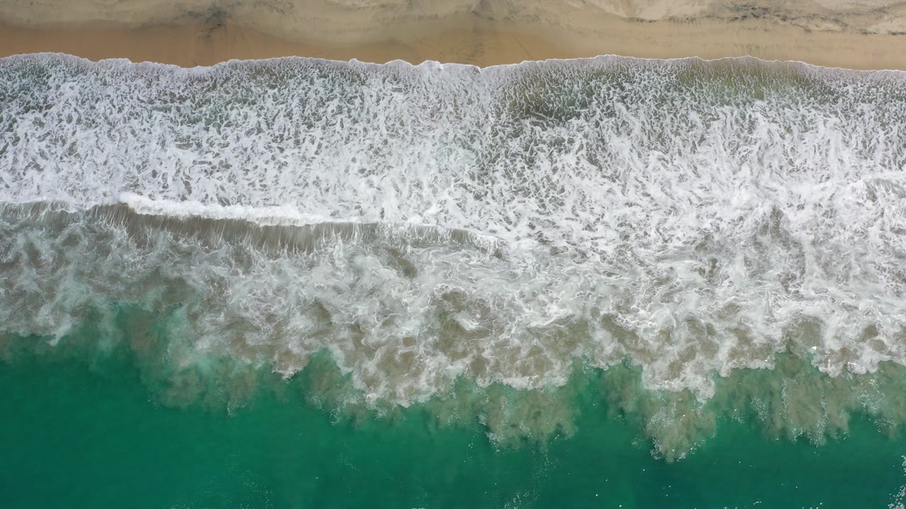 Overhead view of white surf rolling toward tan sand and green-blue water in Los Cabos, Mexico, with no visible people.