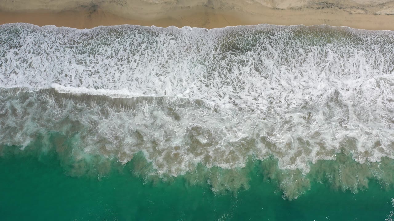 A broad band of white foam spreads across green-blue water below a narrow sandy beach strip in Los Cabos, Mexico, with no visible people.