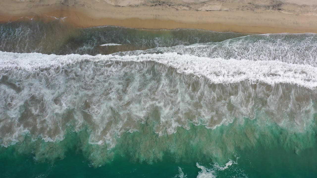 Overhead view of green water and white foam reaching a tan Los Cabos beach, with darker wet sand along the top and no visible people.