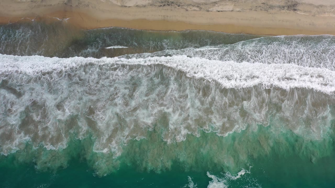 A wide band of whitewater spreads across green Los Cabos surf below a narrow strip of tan beach, with no visible people or structures.