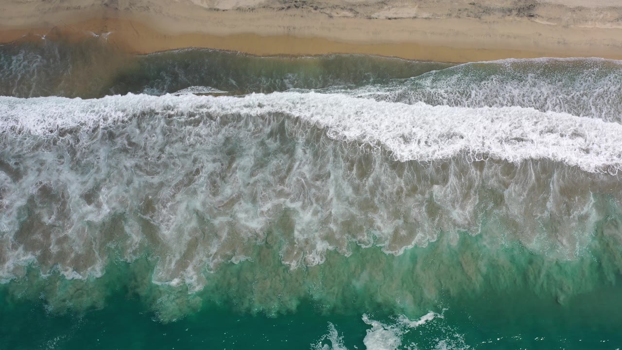 Foamy surf curls in a continuous horizontal line toward the Los Cabos shoreline, with green water below and an empty sand strip above.