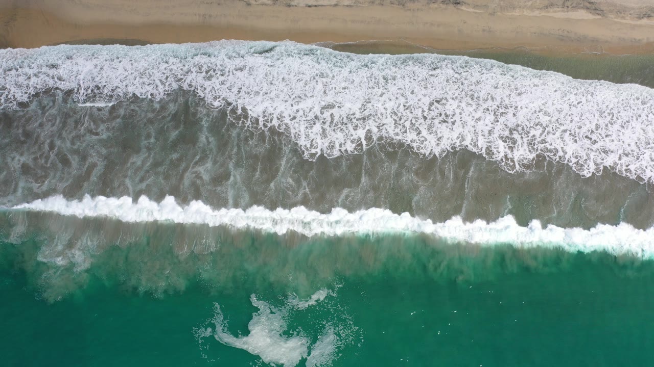 Overhead view of green water and white surf reaching a narrow sand strip in Los Cabos, Mexico, with no visible people.