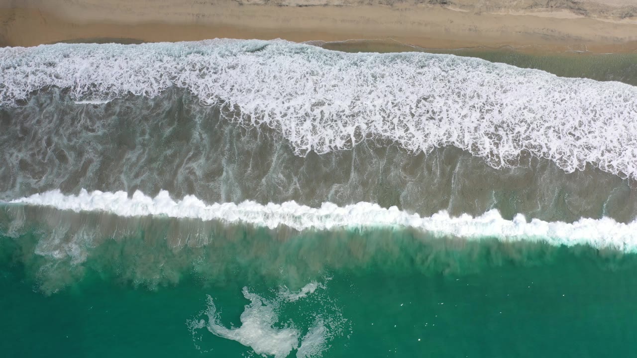 A broad white foam band spreads across green seawater below the sand edge in Los Cabos, Mexico, with no visible people.