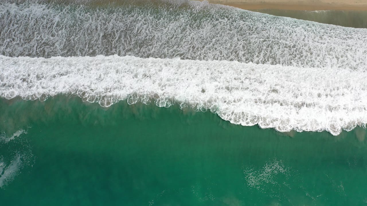 Overhead view in Los Cabos, Mexico, showing a broad band of white surf breaking across green water with dark wet sand at the top and no visible people.
