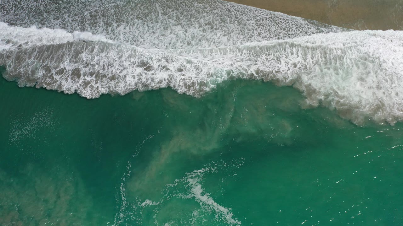 Overhead frame from Los Cabos, Mexico showing white surf along the top edge, teal water below, sandy bottom visible through the water, and no visible people.