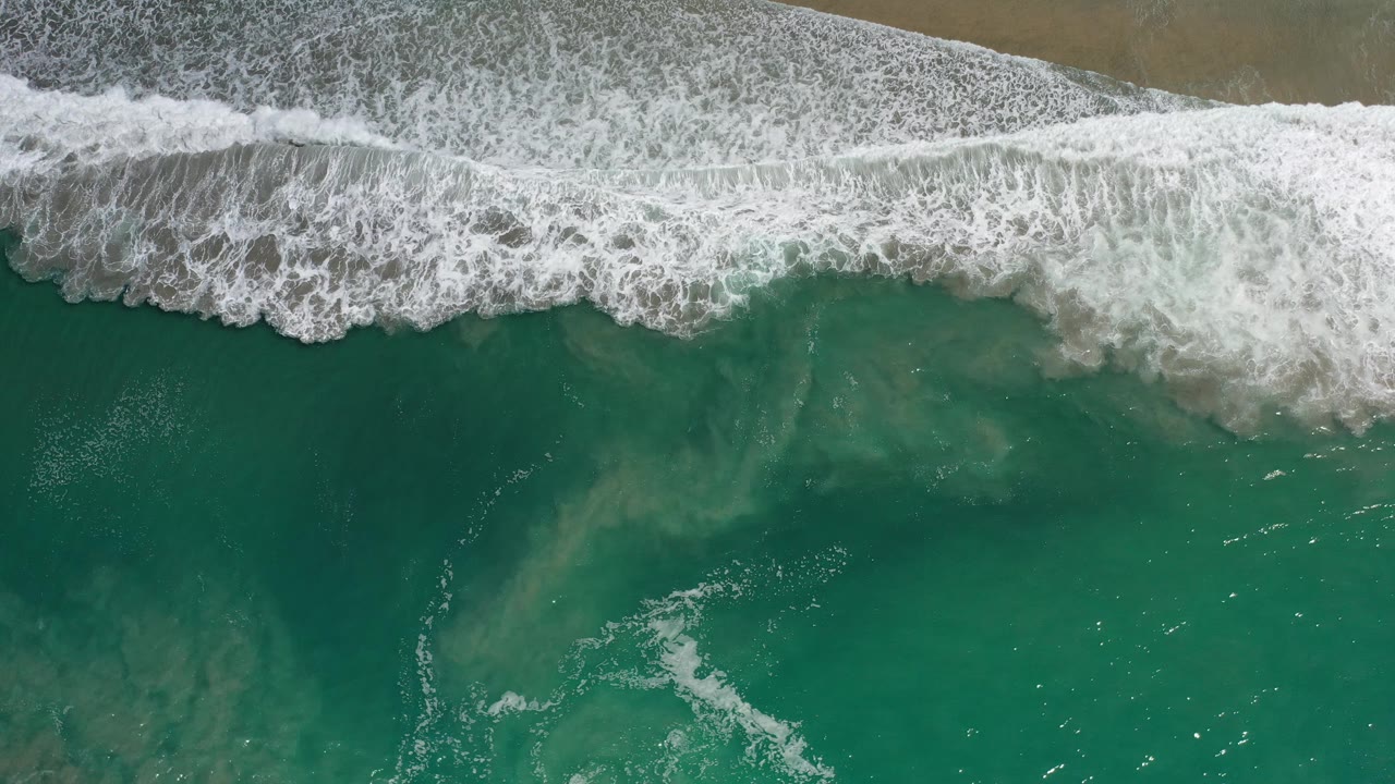 Overhead Los Cabos surf frame with a broad white foam band crossing the upper frame, green-blue water beneath it, faint sand plumes under the surface, and no visible people.