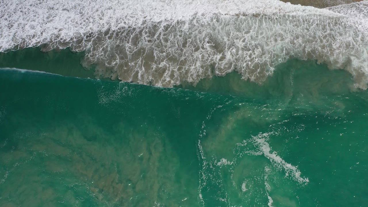 Overhead view of turquoise-green ocean in Los Cabos, Mexico, with a white band of surf foam crossing the top of the frame and no visible people.