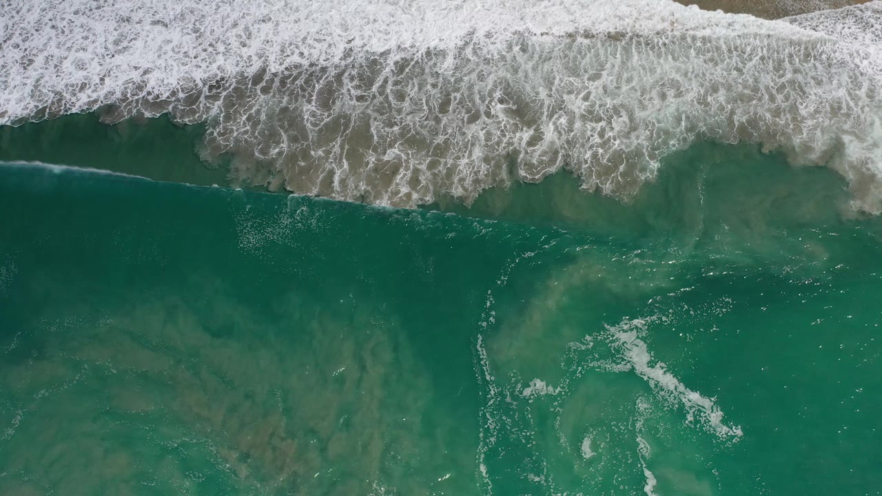 Aerial view over Los Cabos ocean water, where white foam spreads along the upper shoreline band above darker submerged sand patterns, with no visible people.