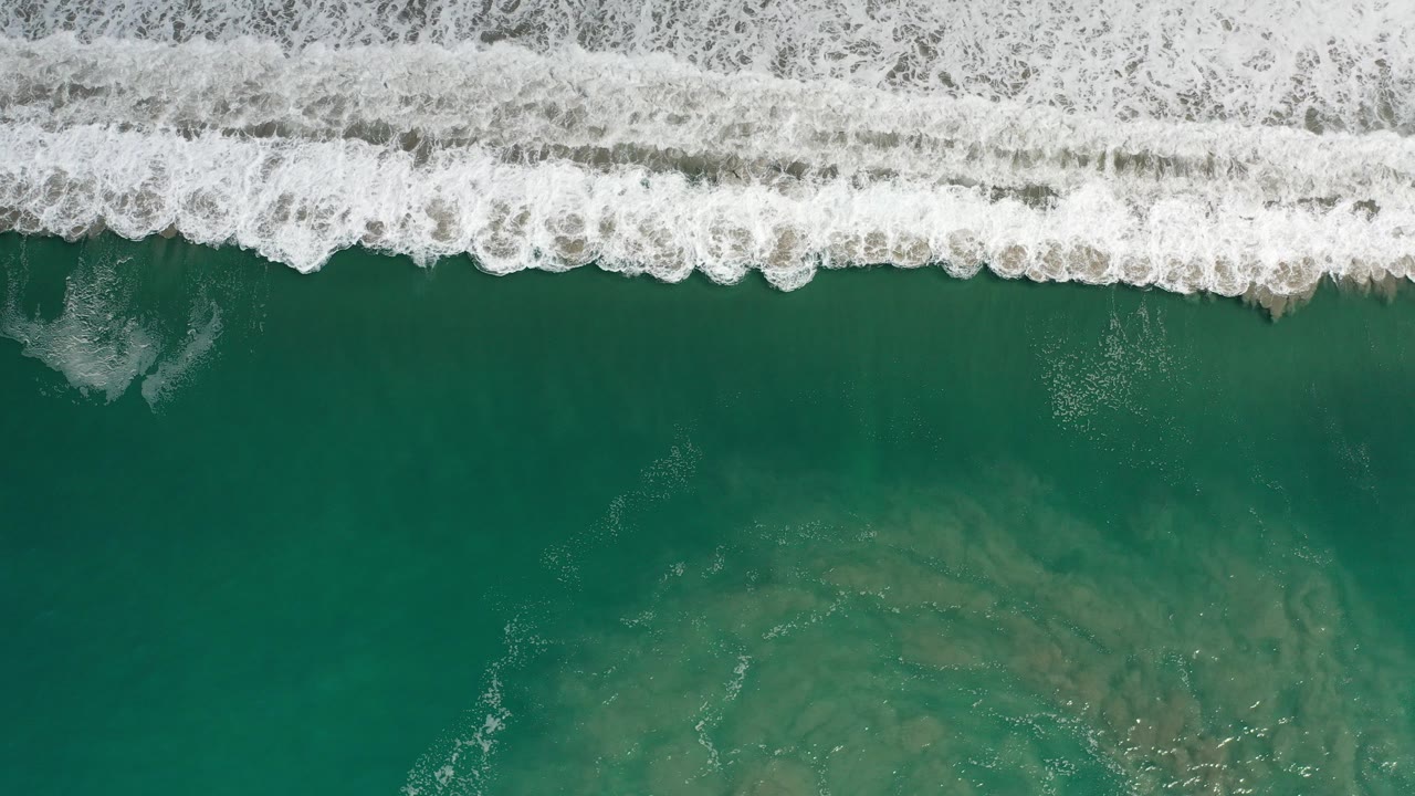 Overhead view in Los Cabos, Mexico, showing a white surf band across the top of green ocean water with faint darker patches below and no visible people.