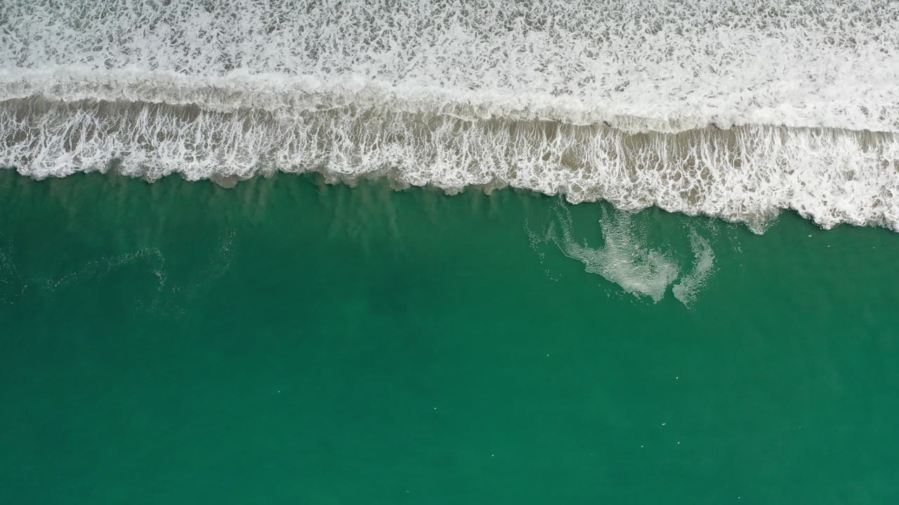 Overhead aerial frame of waves breaking across green water along a sandy Los Cabos shoreline, with no visible people.
