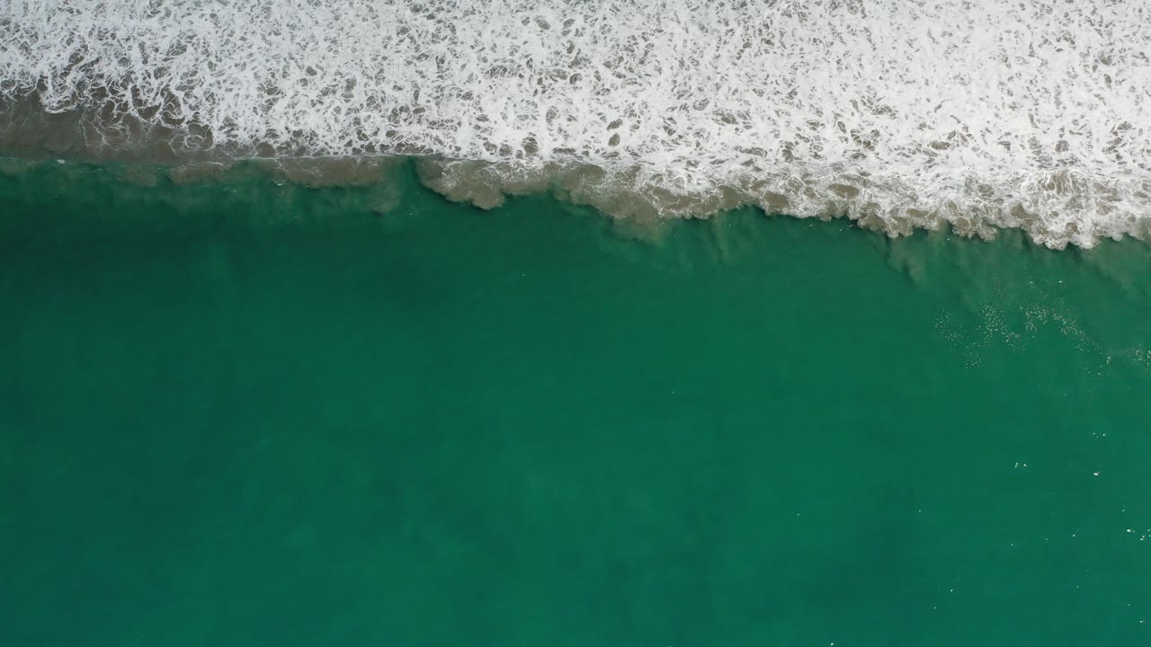 Overhead view of Los Cabos ocean water with a wide band of white surf at the top, darker underwater patches below, and no visible people.