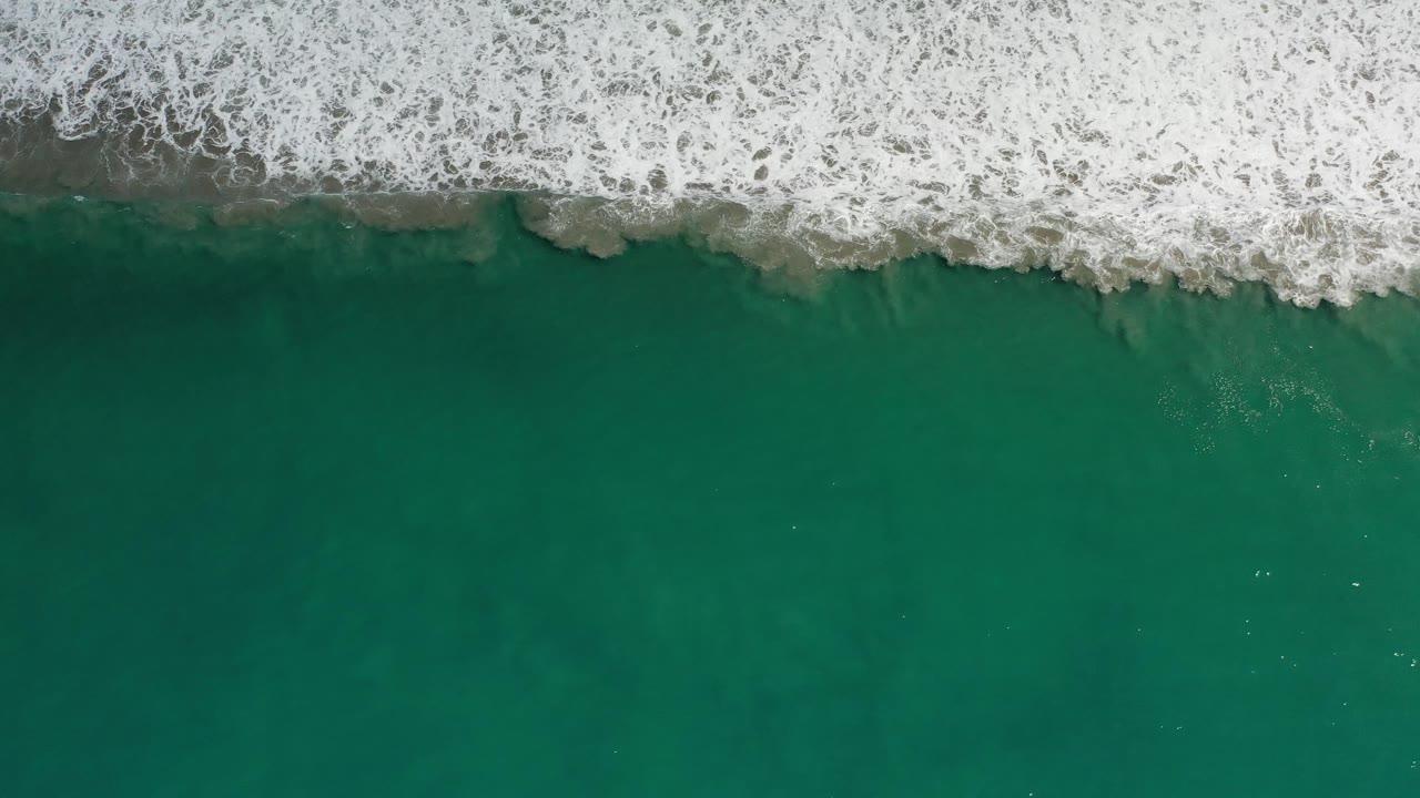Los Cabos surf spreads in a bright white band across the upper frame while green-blue water and faint underwater shapes fill the lower area, with no visible people.