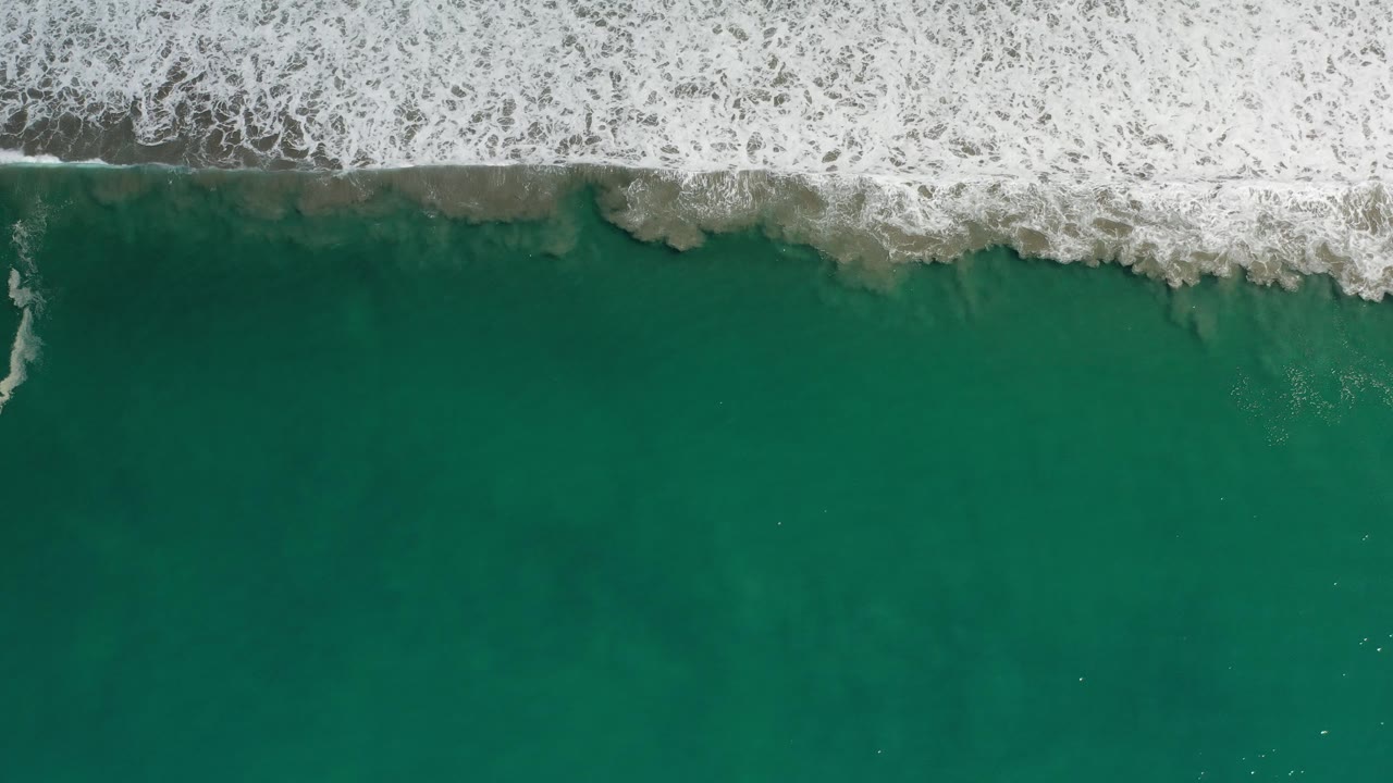 Aerial view of foamy surf above dark teal coastal water.