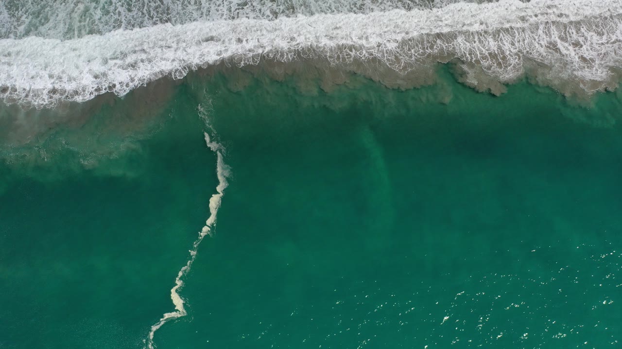 Overhead view of teal water and a white breaking wave along the upper edge in Los Cabos, Mexico, with a thin foam streak crossing the water and no visible people.