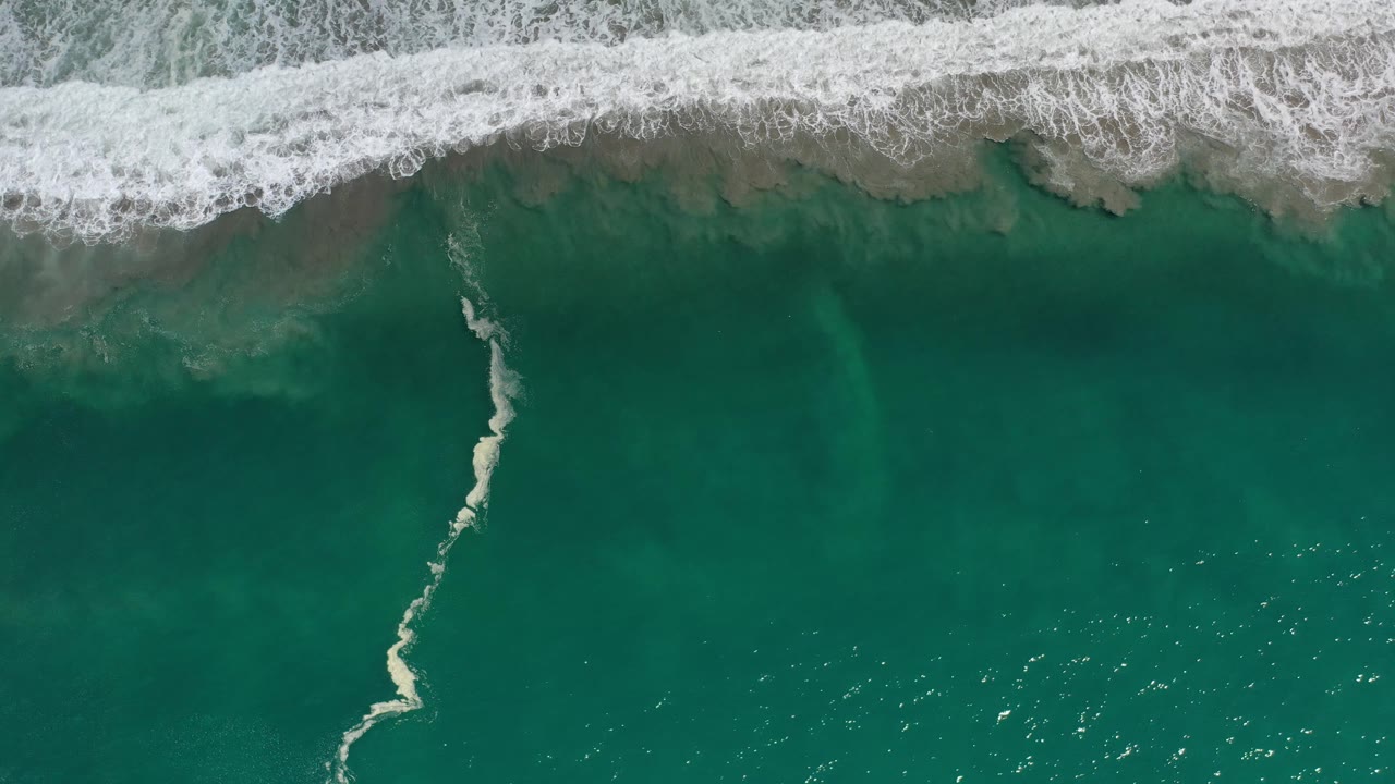 Top-down Los Cabos ocean frame with white foam spreading across the top, green-blue water below, tan sand visible under the surface, and no visible people.