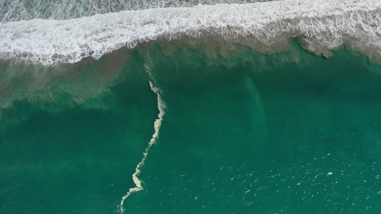 Aerial surf line and dark teal water with a pale foam trail.