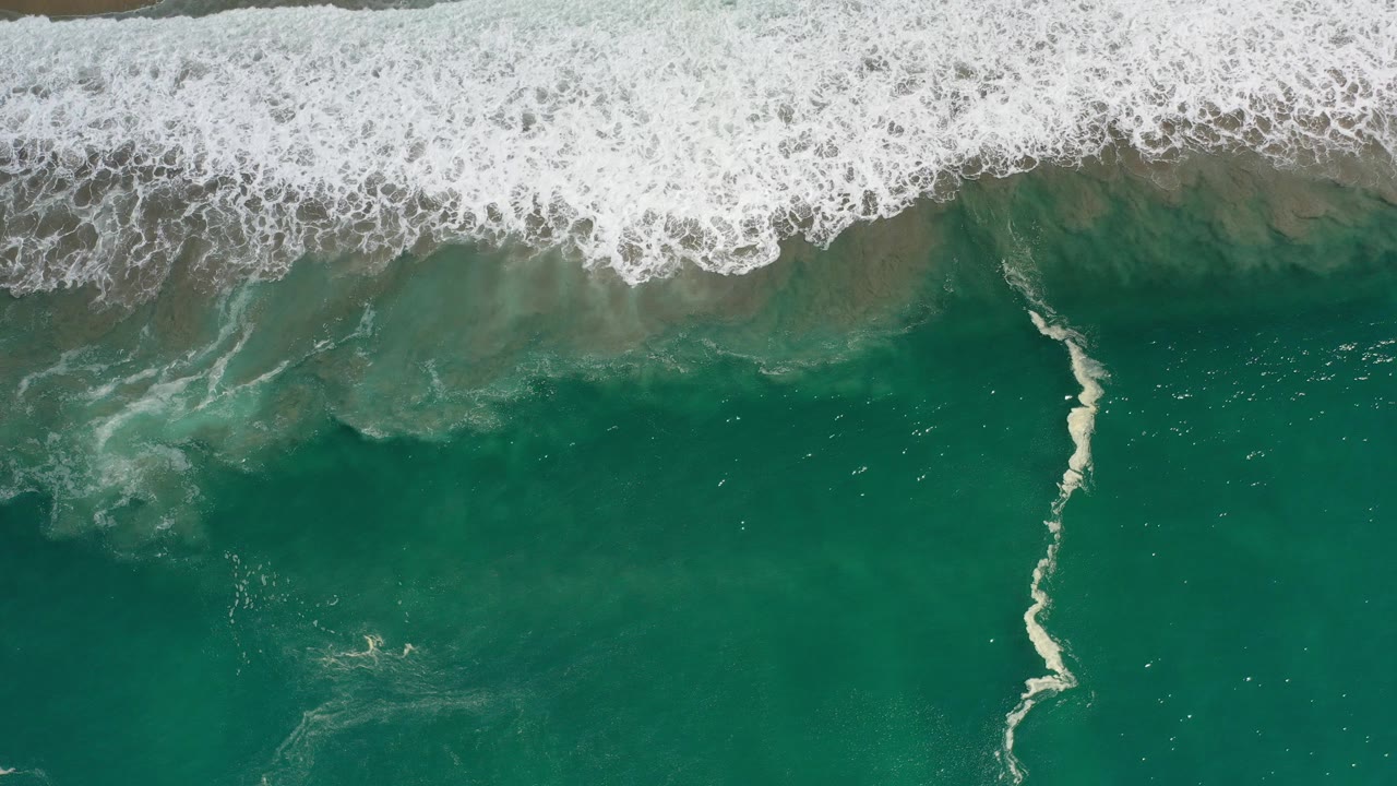 Overhead view of green Los Cabos ocean water with a broad band of white surf at the top and a small pale swimmer-like figure near the lower left water area.