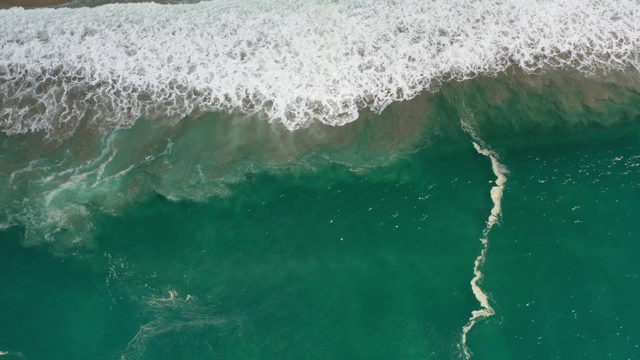 Overhead Los Cabos beach frame with dense white foam along the top, green water below, and a small pale figure visible near the lower center-left.