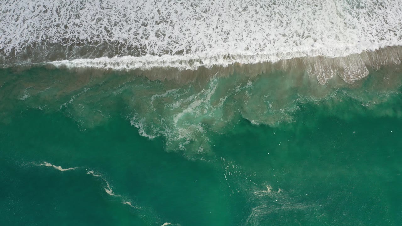 Overhead view of Los Cabos ocean water with a band of white surf at the top, teal water below, and no visible people.