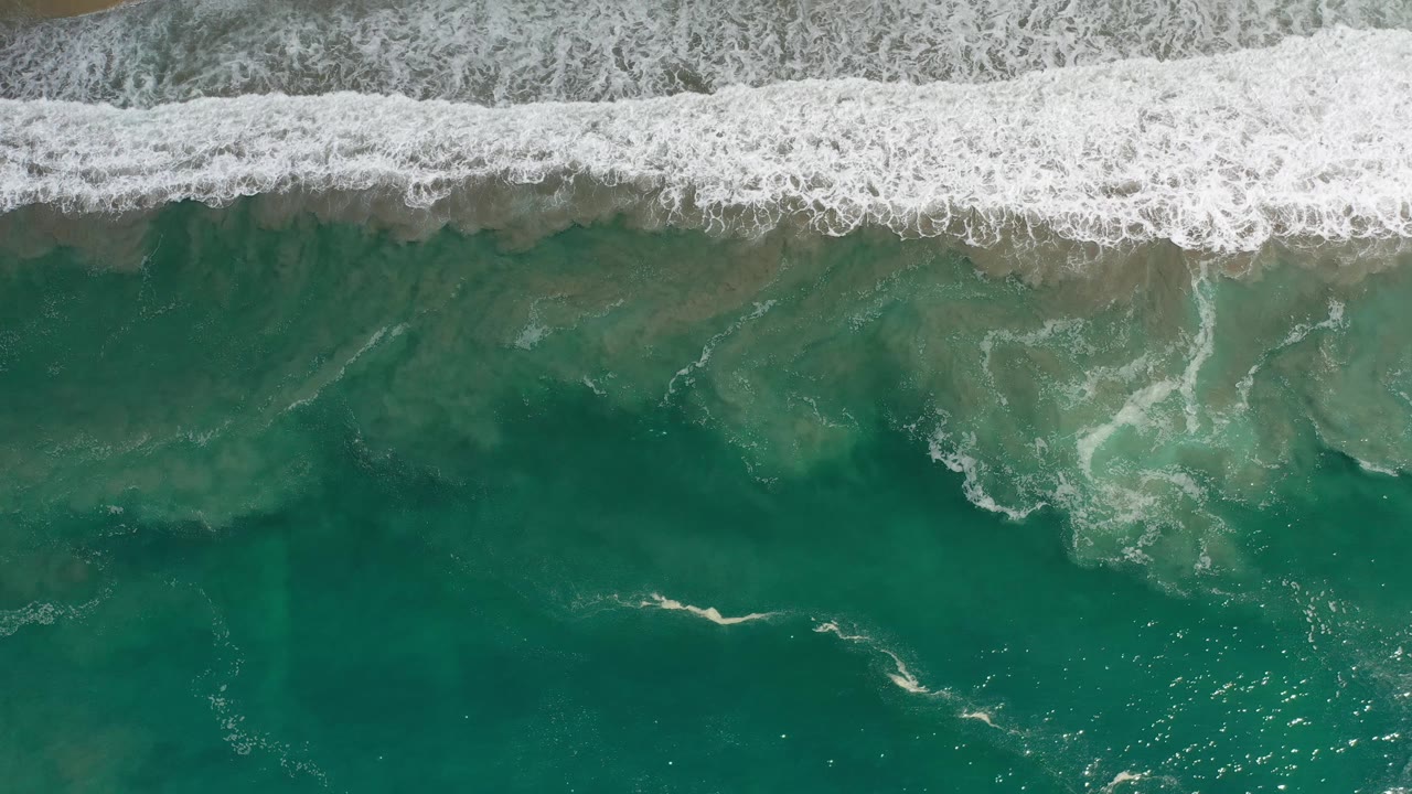 Los Cabos shoreline seen from above as white wave foam stretches horizontally across the frame over green water, with no visible people.