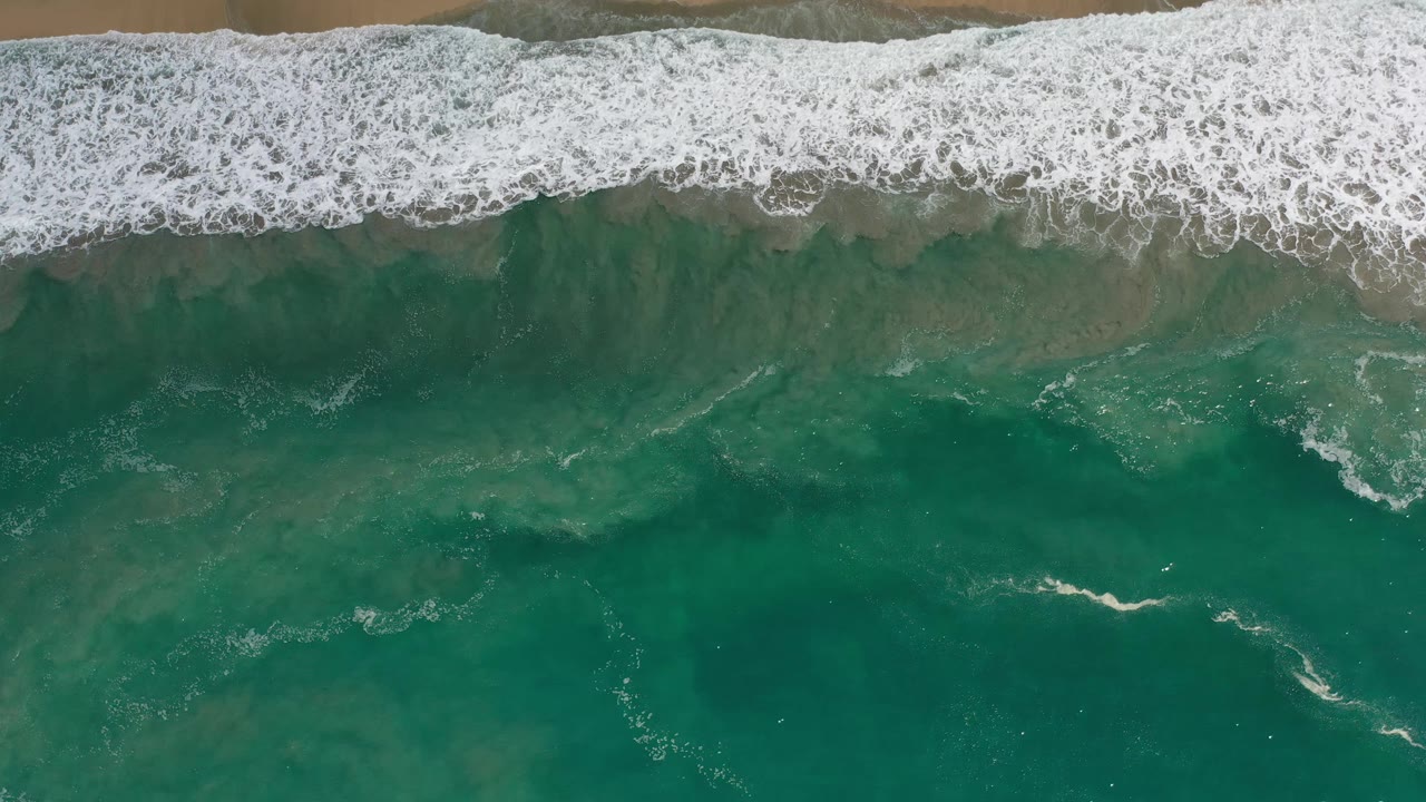 White surf spreads farther across shallow green water near the sand in Los Cabos, Mexico, with no visible people.