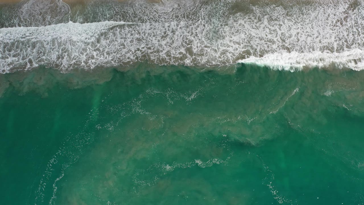 Aerial Los Cabos beach frame with a broad white surf line across the top and green ocean water below, with no visible people.