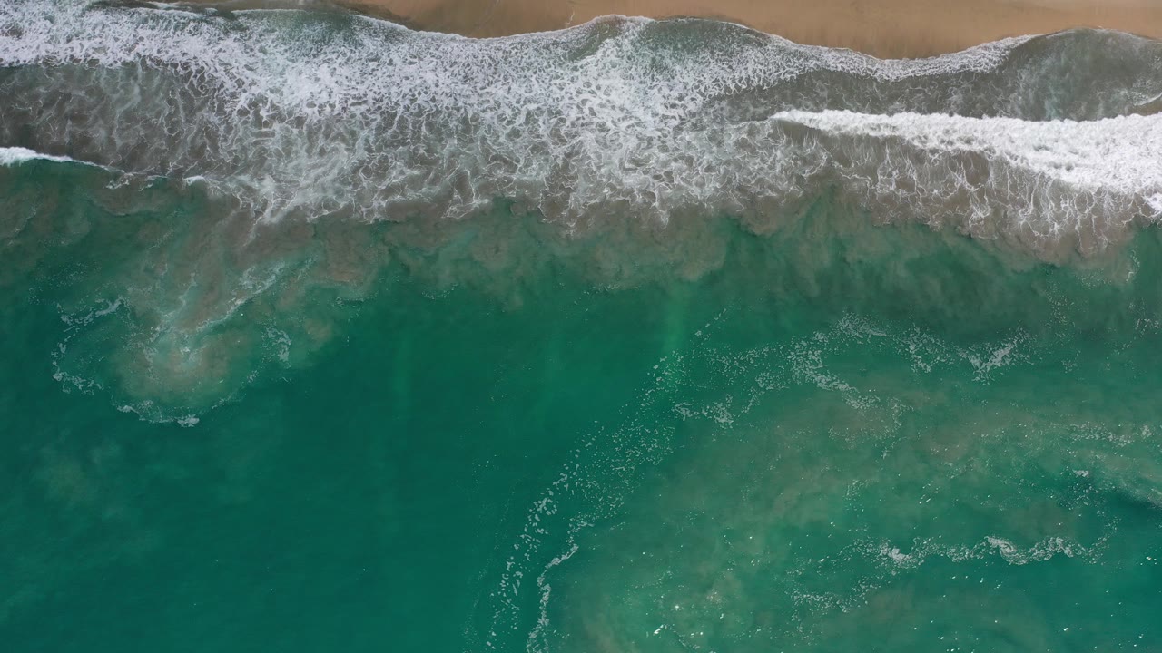 Overhead view of green ocean water and white surf reaching a narrow tan beach in Los Cabos, Mexico, with no visible people.