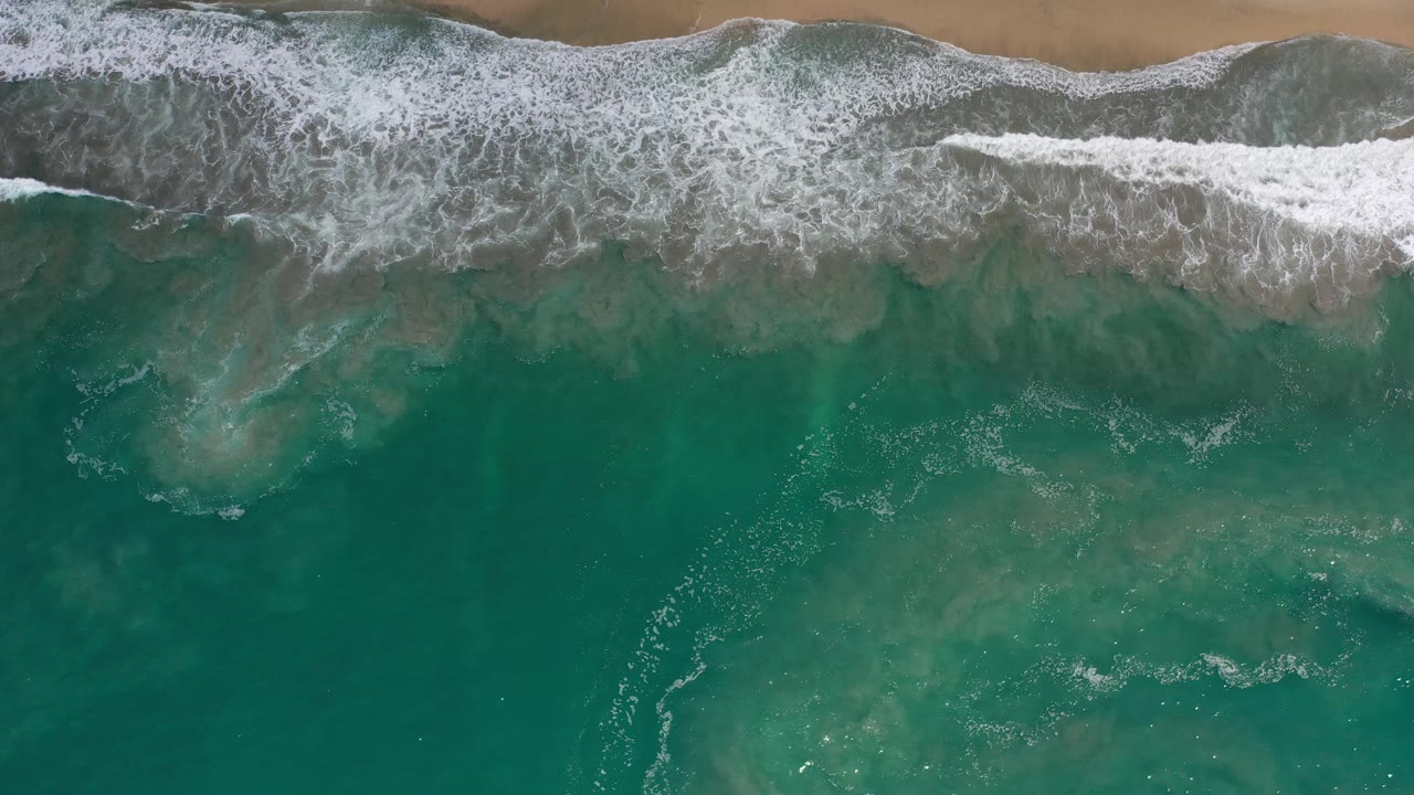 A broad white wave line breaks across teal-green water below the tan beach in Los Cabos, Mexico, with no visible people.