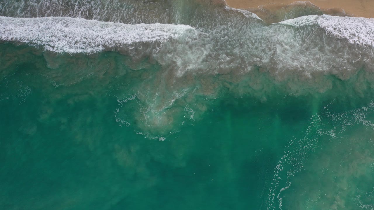 Overhead view of teal ocean and white surf meeting a narrow sandy beach in Los Cabos, Mexico, with no visible people.