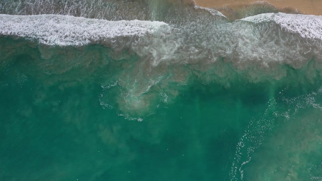A broad band of white foam pushes across green-blue water toward the sand in Los Cabos, Mexico, with no visible people.