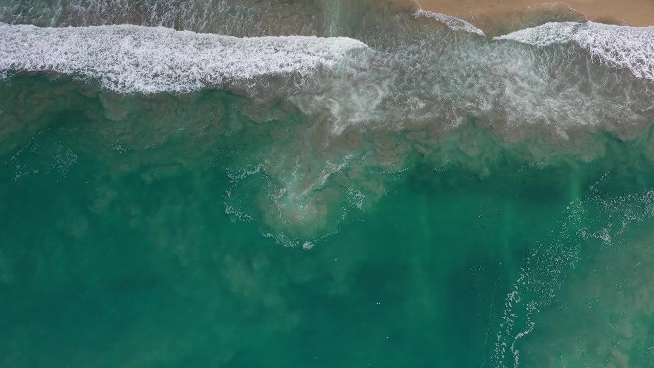 Foamy surf curls along the upper edge of the frame over shallow teal water on the Los Cabos shoreline, with no visible people.