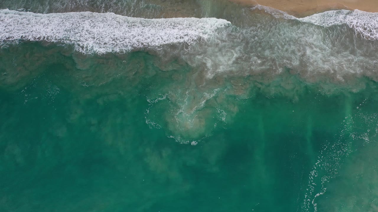 Aerial beach frame with white surf crossing green ocean water.