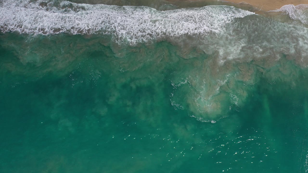 Top-down view of blue-green water and white surf reaching a narrow tan beach in Los Cabos, Mexico, with no visible people.