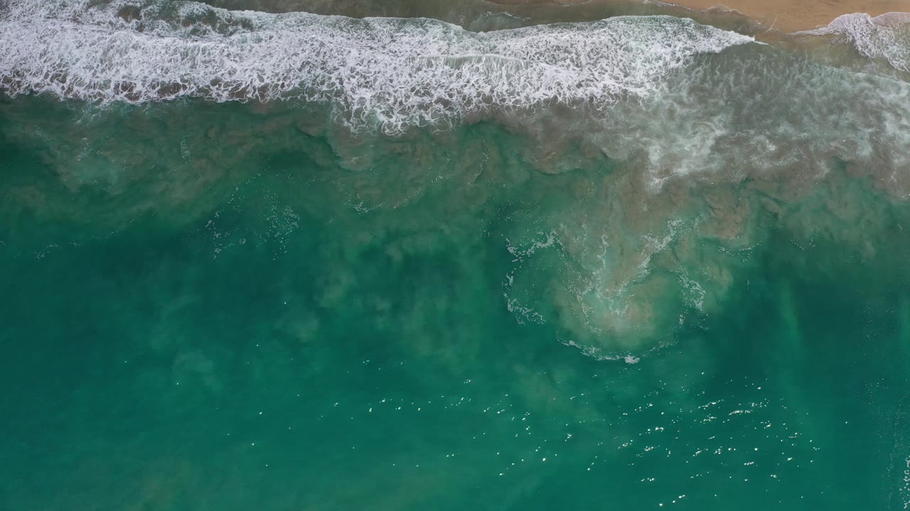 A broad band of foamy surf spreads along the upper edge while darker green water fills the lower frame in Los Cabos, Mexico, with no visible people.