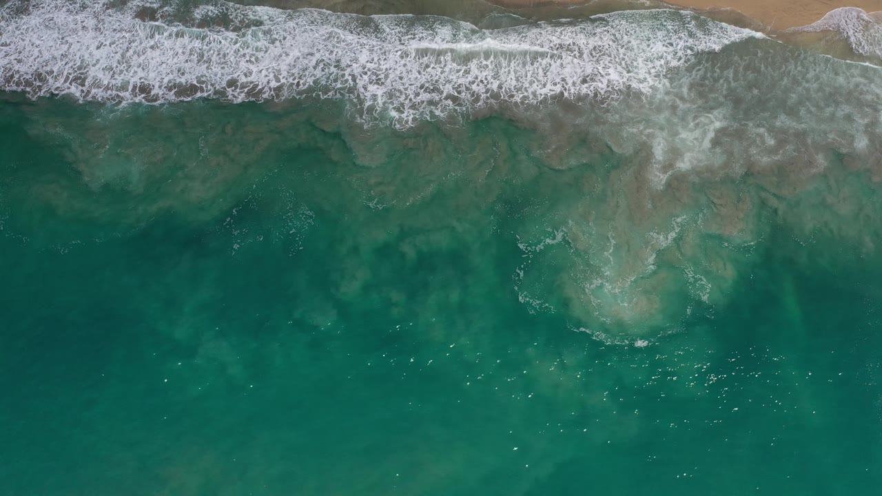 Aerial view of surf breaking across a narrow beach edge.
