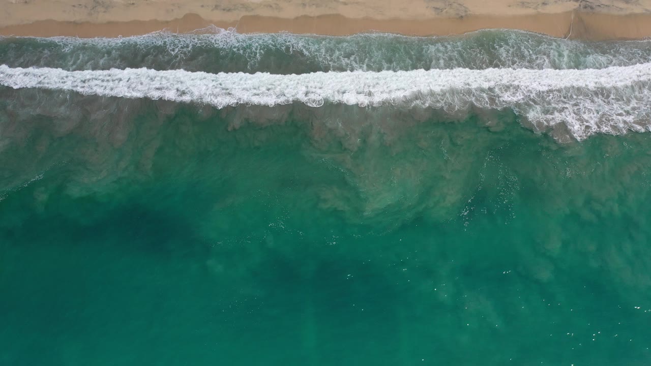 Overhead frame of waves breaking along a sandy Los Cabos beach, with teal water filling the lower frame and no visible people.