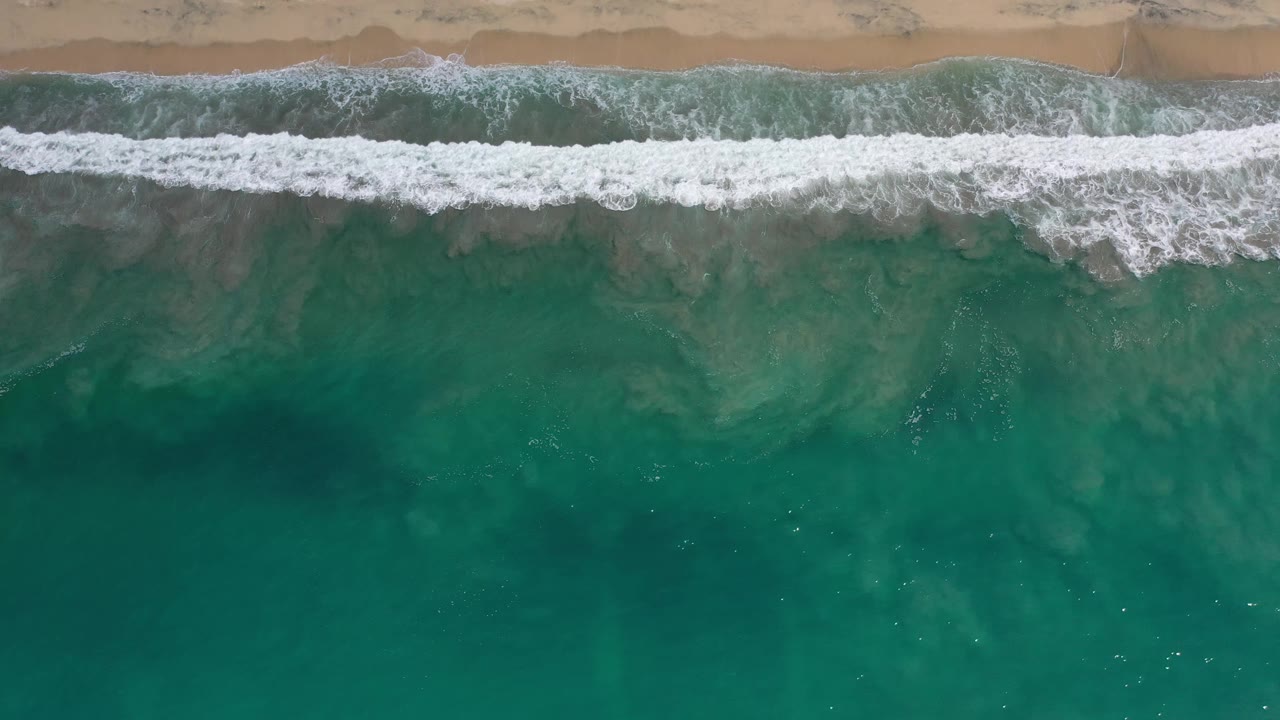 Overhead Los Cabos beach view with a thin tan sand band above a white foam break and green-blue water below, no visible people.