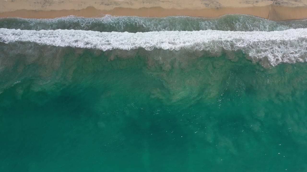 Aerial view of breaking wave foam and darker patches in shallow water.