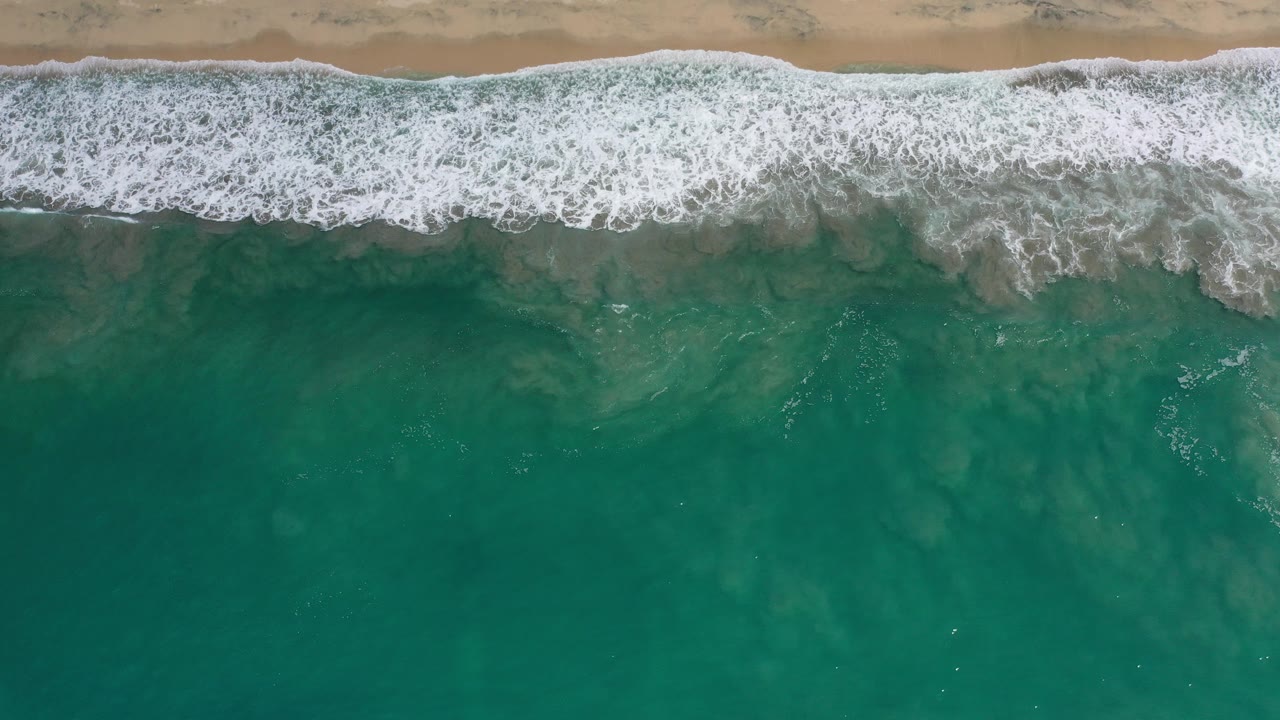 Overhead view in Los Cabos, Mexico of teal ocean water meeting a narrow sandy beach with white foam spread along the shoreline and no visible people.