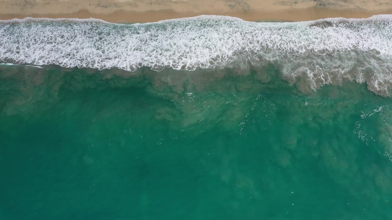 Overhead Los Cabos beach frame with a wider white foam band breaking across tan sand while darker green water fills the lower portion and no visible people.