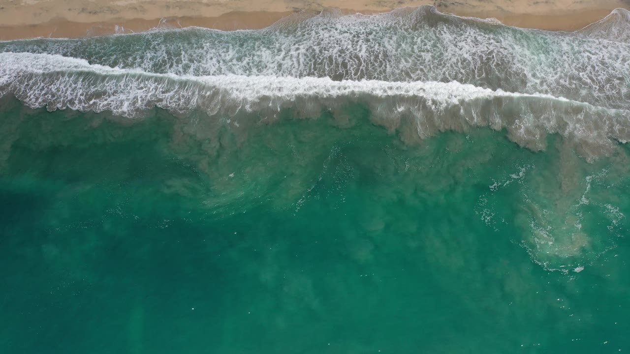A Los Cabos overhead beach frame with foamy surf stretched horizontally below wet sand and deeper green-blue water in the foreground.