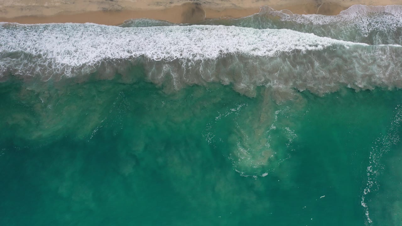 Overhead surf line and turquoise water beside a sandy beach.